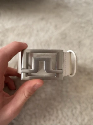 Vitt skärp från J.Lindeberg - Snyggt vitt skärp från J.Lindeberg med bred rem i skinn och en stor, silverfärgad metallspänne med grafisk design. Perfekt för att lyfta vilken outfit som helst med en clean och modern look. Märket är präglat på läderöglan. Längd 110cm. Hör av er vid fler frågor.