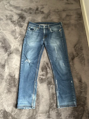 Blå slitna Levi's jeans med hål - Säljer ett par blå Levi's jeans med slitningar och hål på vänster knä och ficka. Jeansen har klassisk femficksmodell, raka ben och ljus tvätt med snyggt sliten look. Perfekta för en avslappnad och trendig stil.