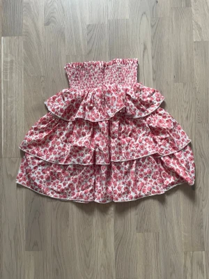 Blommig volangkjol med hög midja - Söt blommig kjol med flera volanger och hög smockad midja. Kjolen har ett rosa och rött blommönster på vit botten och är perfekt för sommarens alla tillfällen. Tillverkad i lätt bomullstyg som ger en luftig känsla.