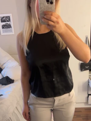 Svart satinlinnetopp med korsade band - Snygg svart linnetopp i satin med tunna axelband som korsas i ryggen. Toppen är ärmlös och har en öppen rygg. Perfekt att matcha med både jeans och kjol.