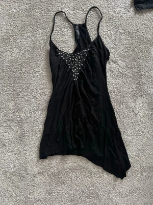 Svart linnetopp med pärldetaljer - Snygg svart linnetopp med tunna axelband och racerback. Toppen har dekorativa pärlor i triangelform framtill som ger en extra touch. Asymmetrisk nederkant för en cool look. Perfekt att styla med jeans eller kjol.💗💗