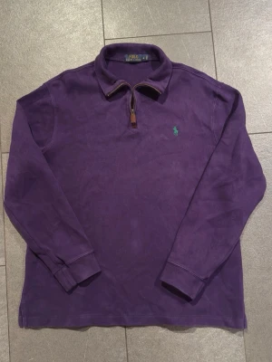 Lila halvzip från Polo Ralph Lauren - Säljer en lila långärmad halvzip från Polo Ralph Lauren med klassisk krage och dragkedja med läderdetalj. Tröjan har det ikoniska gröna Polo-märket broderat på bröstet och är tillverkad i mjukt bomullsmaterial.