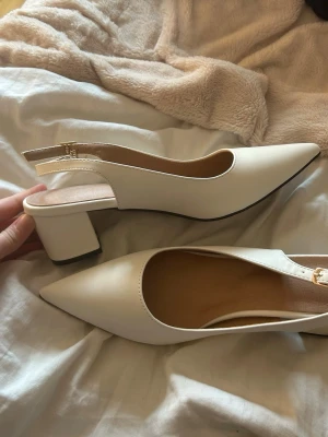 Vita slingback pumps med blockklack - Stilrena vita pumps med spetsig tå och öppen häl. Skorna har en bred blockklack och justerbart slingback-band med guldfärgat spänne. Tillverkade i slätt konstläder för en modern look.
