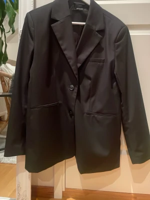 Svart kavaj från Gina Tricot - Stilren svart kavaj från Gina Tricot med klassisk krage och två knappar framtill. Kavajen har två fickor och en bröstficka, samt en rak passform som ger en snygg siluett. Perfekt att styla till olika looks.