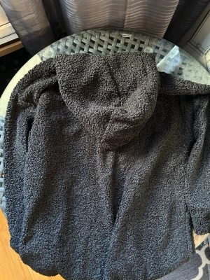 Svart teddytröja med huva - Mysig svart teddytröja med huva i fluffigt material. Tröjan har en avslappnad passform och är perfekt för att hålla värmen under kyliga dagar. Inga synliga märken eller detaljer på framsidan. Passar dig som gillar en enkel och stilren look.