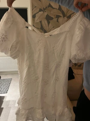 Vit off shoulder klänning med spets - Söt vit klänning med off shoulder-modell och broderade blommor. Klänningen har puffiga korta ärmar, spetsdetaljer längs kanterna och volang nertill. Knytband i ryggen ger en extra fin detalj. Perfekt för sommarens alla tillfällen. Helt oanvänd då och ny pris är 650kr
