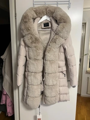 Ljusbeige dunjacka med fuskpäls - Säljer en lång, ljusbeige dunjacka från Cappuccino med stor fluffig fuskpäls runt huvan och längs dragkedjan. Jackan har bälte i midjan och två fickor med dragkedja. Perfekt för kalla vinterdagar och riktigt snygg design.