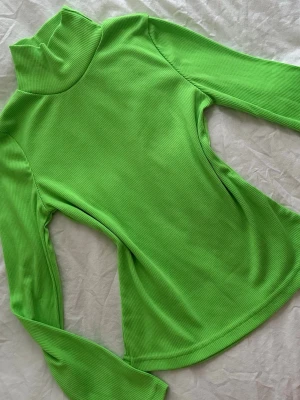 Limegrön ribbad polotröja - Säljer en limegrön ribbad polotröja med lång ärm och hög krage. Tröjan är figurnära och har ett stretchigt material som ger en snygg passform. Perfekt för dig som gillar starka färger och vill sticka ut.