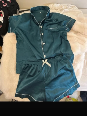 pyjamas set i petrolblå - Snyggt pyjamasset i glansigt petrolblått med kortärmad skjorta och matchande shorts. Skjortan har vita knappar, kontrasterande vita kanter och en bröstficka. Shortsen har en söt vit rosett i midjan och vita detaljer längs bensluten. Passar storlek S-M-L