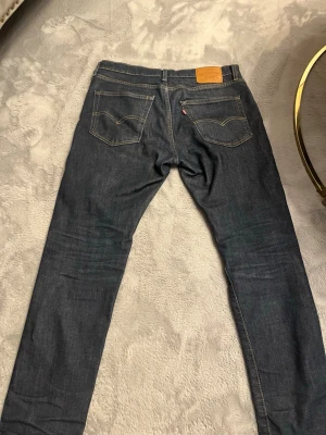 Mörkblå Levi's jeans med raka ben - Säljer ett par klassiska mörkblå jeans från Levi's med raka ben och kontrastsömmar. Jeansen har två bakfickor med den ikoniska Levi's-sömmen och en liten röd etikett på fickan. Tillverkade i slitstark denim, perfekta för en avslappnad och cool stil.