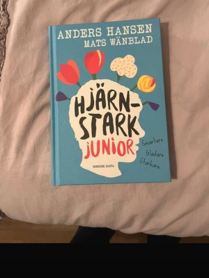 Hjärnstark junior av Hansen & Wänblad - En färgglad bok med blått hårt omslag, skriven av Anders Hansen och Mats Wänblad. Titeln 'Hjärnstark junior' står i svart och rött, med illustrationer av blommor och rullskridskor. Perfekt för unga som vill bli smartare, gladare och starkare.