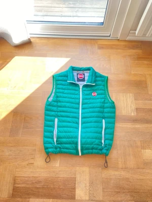 Grön dunväst från Colmar - Snygg grön dunväst från Colmar med vit dragkedja och två fickor framtill. Västen har en hög krage, quiltad design och Colmar-logga på bröstet. Perfekt för lager-på-lager och enkel att matcha med olika outfits.