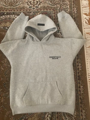 Essentials tracksuit - Säljer ett grått loungewear-set från Fear of God Essentials. Setet består av en hoodie med magficka och tryck på bröstet samt matchande mjukisbyxor med dragsko och tryck på benet. Perfekt för chill dagar hemma eller när du vill vara bekväm.