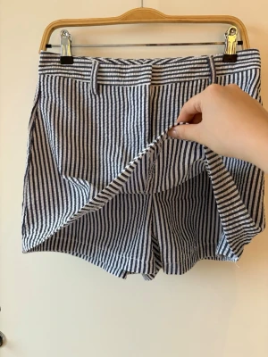 Randig skort med blåvita ränder - Säljer en snygg skort med blå och vita vertikala ränder. Modellen har bälteshällor, fickor och en omlottfront som ger kjol-look fram och shorts bak. Perfekt för dig som vill ha både stil och komfort. Materialet känns lätt och luftigt, troligen bomull eller linne. Endast testad, aldrig använd, väldigt liten i strl så passar ngn med strl S. linen shorts, striped shorts women, skort, blue white striped, high waisted shorts, summer shorts, coastal style, european summer outfit, vacation outfit, ligh