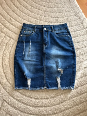 Blå jeanskjol med slitningar - Säljer en blå jeanskjol i kort modell med slitna detaljer och fransad nederkant. Kjolen har fem fickor, bälteshällor och knappstängning fram. Perfekt för dig som gillar en avslappnad och trendig stil.