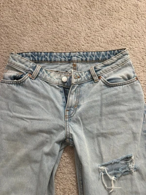 Ljusblå raka jeans med slitningar - Säljer ett par ljusblå jeans med raka ben och slitningar på ena låret. Jeansen har klassisk femficksmodell, råa kanter vid benslut och coolt mönstrat innerfoder i midjan. Perfekta för en avslappnad och trendig look.