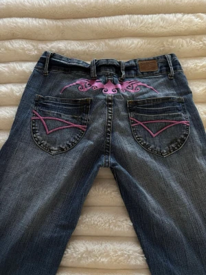 Blå jeans med rosa brodyr Crazy Age - Unika blå jeans från Crazy Age med rosa broderade detaljer på bakfickorna och ovanför fickorna. Klassisk femficksmodell med raka ben och slitningar för en cool look. Materialet är denim i bomull med stretch för extra komfort. Mått: 36cm rakt över i midjan, innerbenslängd 73cm (avklippta)
