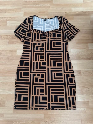 Ny. Beige och svart mönstrad klänning - Kortärmad klänning med beige och svart grafiskt rutmönster. Klänningen har en rak passform och fyrkantig ringning. Tillverkad i mjukt och stretchigt material, perfekt för dig som gillar statement-plagg. Storlek xl, men förminskad som m/l