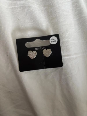 Två par silvriga örhängen - Säljer två par örhängen. Ett par från Montini i form av glittriga hjärtan i silverfärg och ett par från Ur & Penn med runda, klara stenar som ger en elegant look. Båda paren är små och stilrena, perfekta för att lyfta vilken outfit som helst. Aldrig använt dem!!