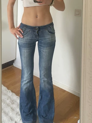 Blå bootcut jeans med låg midja - Säljer ett par blå bootcut jeans med låg midja och slitna detaljer framtill. Jeansen har klassiska fem fickor och stängs med dragkedja och knapp. Perfekta för en avslappnad och trendig look.