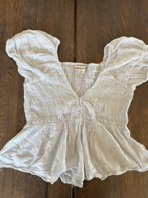Vit blus från Urban Outfitters - Supersöt vit blus från Urban Outfitters med puffärmar och v-ringning. Toppen har smockad midja och lätt volang nedtill för en feminin look. Perfekt till jeans eller kjol och gjord i ett tunt, luftigt bomullstyg.