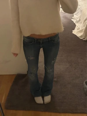 Blå slitna bootcut jeans - Säljer ett par blå bootcut jeans med slitningar på låren och knäna. Jeansen har låg midja och klassisk femficksdesign. Perfekta för en avslappnad och trendig look. Materialet är denim i bomull.