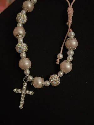 Pärlhalsband med glittrigt kors - Snyggt halsband med stora ljusrosa pärlor, mindre vita pärlor och glittrande kulor. Halsbandet har ett hängsmycke i form av ett kors dekorerat med strass. Trädd på rosa snöre med justerbar knut. Perfekt för dig som gillar bling och statement-smycken.