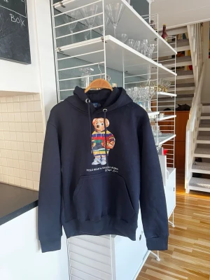 Marin blå  Polo Bear Ralph Lauren - Svart hoodie från Polo Ralph Lauren med den ikoniska Polo Bear-figuren på bröstet i färgglada kläder. Klassisk känguruficka framtill, justerbar huva med snören och ribbade muddar. Perfekt för en avslappnad och trendig stil. Helt ny