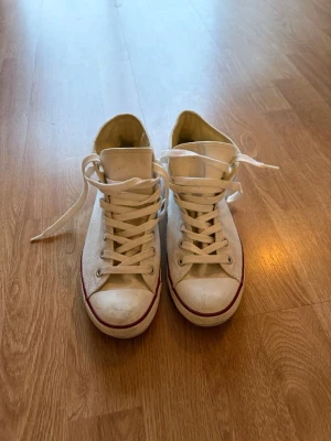 Vita Converse Chuck Taylor Highs - Klassiska vita Converse Chuck Taylor All Star high tops med vit canvas, vita snören och gummisula med röd rand. Ikonisk logga på sidan och metallöljetter för snörningen. Perfekta för en avslappnad och tidlös stil.