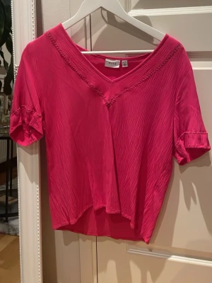 Rosa v-ringad topp från Vila i strl 40 - Snygg rosa topp från Vila med v-ringning och korta ärmar. Toppen har en lätt glansig yta och dekorativa sömmar längs halsringningen. Perfekt att styla med jeans eller kjol för en trendig look.