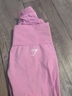 Rosa tights från Gymshark - Säljer ett par rosa tights från Gymshark med hög midja och diskret mönster längs benen. Tightsen har en tight passform och är perfekta för gymmet eller annan träning. Tillverkade i stretchigt material för maximal komfort. Använd ett fåtal gånger. Hör av dig om du har frågor🥰