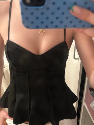 Svart korsettopp med tunna axelband - Snygg svart korsettopp med tunna axelband och markerad midja. Toppen har en lätt utsvängd nederdel och en figurnära passform som framhäver siluetten. Perfekt att styla med jeans eller kjol för en trendig look.