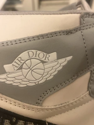 Air Dior x Nike gråvita sneakers - Exklusiva Air Dior x Nike sneakers i grått och vitt läder med ikonisk Air Dior-logga på sidan och genomskinlig sula med Dior-tryck undertill. Snygg och unik design som sticker ut, perfekt för dig som gillar streetwear och high fashion.