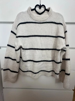 Vit stickad tröja med svarta ränder - Säljer en vit stickad tröja med svarta horisontella ränder och ribbad krage. Tröjan har en avslappnad passform och långa ärmar, perfekt för en chill och stilren look. Materialet känns mjukt och mysigt, perfekt för kyligare dagar. 