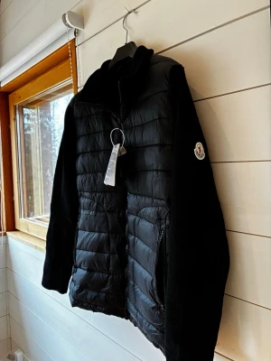 Svart pufferjacka från Moncler - Snygg svart pufferjacka från Moncler med quiltad framsida, dragkedja och Moncler-logga på ärmen. Jackan har höga krage och stickade ärmar och rygg för extra komfort. Perfekt för kalla dagar och har praktiska fickor med dragkedja.