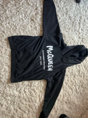 Svart hoodie Alexander McQueen - Snygg svart hoodie från Alexander McQueen med stor vit logga och texten 'EST. 1992' på bröstet. Klassisk känguruficka framtill och huva med dragsko. Perfekt för en streetwear-look och riktigt bekväm att bära.