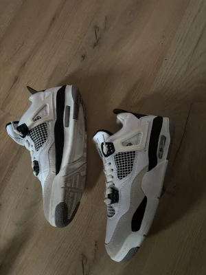 Vita och svarta Air Jordan 4 sneakers - Säljer ett par Air Jordan 4 sneakers i vitt och svart med klassisk meshpanel på sidorna, synlig Air-dämpning i sulan och detaljer i plast. Skorna har en robust design och ikoniska vingar vid snörningen. Perfekt för dig som gillar streetstyle.