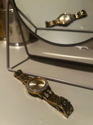 Guldig klocka med stenar - Snygg guldig klocka med metallarmband och glittrande stenar runt urtavlan. Urtavlan är enkel och stilren i guldton, vilket ger en lyxig känsla. Perfekt accessoar för att lyfta din outfit. Nypris 499kr