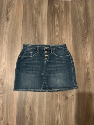 Mörkblå jeanskjol med knappar - Snygg mörkblå jeanskjol med fyra silverfärgade knappar framtill och klassiska fickor både fram och bak. Kjolen har en kort passform och är tillverkad i denim med en lätt tvättad look. Perfekt att matcha med en enkel t-shirt eller hoodie.