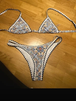 Mönstrad trekantsbikini i vitt och blått - Säljer en snygg trekantsbikini med orientaliskt mönster i vitt, blått och beige. Bikinin har smala axelband och hög skärning på underdelen. Materialet är stretchigt och bekvämt, perfekt för stranden eller poolen. Inga synliga loggor. Osäker på storleken men skulle säga S/M/L beroende hur man vill att den ska sitta