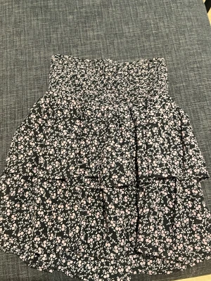 Svart blommig volangkjol - Supersöt kort kjol med svart botten och små blommor i vitt och rosa. Kjolen har bred smock i midjan och dubbla volanger för extra flowy känsla. 