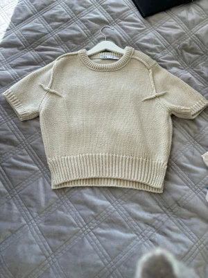 Beige stickad t-shirt från Zara - Kortärmad stickad t-shirt i beige från Zara. Tröjan har en rak passform, ribbstickade kanter vid ärmslut och nederkant samt markerade sömmar vid axlarna. Perfekt för en avslappnad och trendig look.