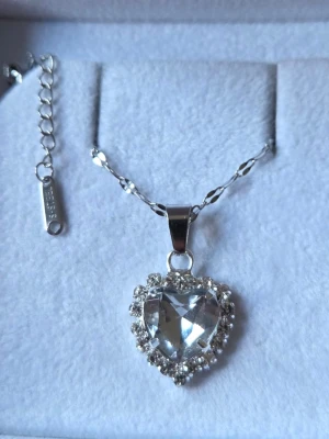 Halsband med hjärthänge  - Snyggt halsband i silverfärg med ett stort hjärtformat hänge i klar kristall. Hänget är dekorerat med gnistrande små stenar runt om och kedjan har en elegant tvinnad design. Perfekt accessoar för att lyfta din outfit. Kedjan är i rostfritt stål (tål vatten & tappar inte sin färg).