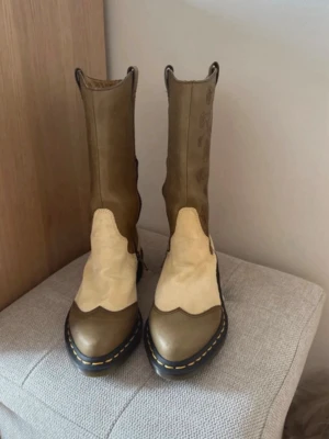 Bruna och beige cowboy boots - Säljer ett par snygga cowboy boots i brunt och beige med blommigt mönster på skaftet. Skorna har kontrastsömmar och robust sula med gul detalj. Tillverkade i läder och mocka, perfekta för dig som vill sticka ut med westernstil.