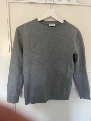 Grå stickad tröja från Kappahl - En enkel och stilren grå stickad tröja från Kappahl. Tröjan har rund halsringning, långa ärmar och ribbade muddar vid ärmslut och nederkant. Perfekt basplagg i garderoben och passar till det mesta. Materialet ser ut att vara mjukt och bekvämt.