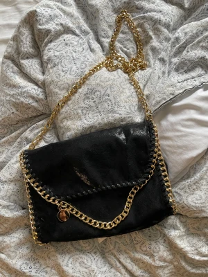 Svart axelväska med guldkedja - Snygg svart axelväska med kraftig guldfärgad kedja och dekorativ kedjedetalj längs kanten. Väskan har ett lock med sömmar och en rund guldfärgad berlock. Perfekt för dig som vill ha en trendig och stilren accessoar till din outfit.
