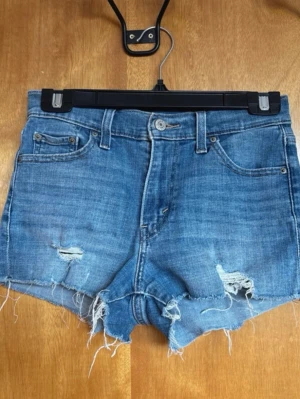 Klassiska Levi’s shorts i perfekt ljusblå tvätt – Storlek 25  - Säljer mina snygga Levi’s shorts som har den där helt rätta vintage-känslan. De är i en klassisk ljusblå färg med snygga slitningar framtill och råa, fransade kanter nedtill som ger en perfekt cut-off look. Shortsen är i jättefint skick och den klassiska läderlappen och bågsömmarna på bakfickorna är helt intakta. Storleken är W25, vilket motsvarar en XS eller en liten 34. De är verkligen de ultimata sommarshortsen som passar till allt från en enkel t-shirt till en finare blus. 