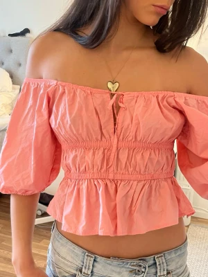 Rosa offshoulder topp från H&M korall - Supersöt rosa offshoulder topp från H&M med puffiga trekvartsärmar och smock i midjan. Toppen har volang nedtill och en liten knytning framtill. Perfekt för en trendig och somrig look.