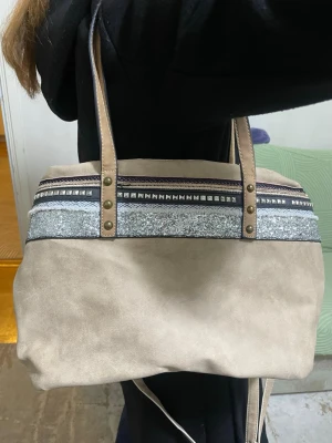 Beige handväska - Snygg och rymlig handväska i beige/neutral färg med fina detaljer som glitterband och nitar. perfekt som vardagsväska eller till resor. Väskan har både handtag och en längre justerbar axelrem, så du kan bära den som handväska eller crossbody. Den är rymlig och har flera fack inuti, vilket gör den praktisk för allt du behöver under dagen.  Viss missfärgning finns på undersidan av väskan (se bild), men inget som syns särskilt när väskan används. I övrigt hel, fräsch och fullt funktionell. 