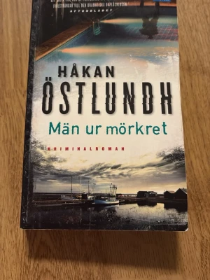 Män ur mörkret av Håkan Östlundh - En spännande kriminalroman av Håkan Östlundh med omslag i blå, svart och vit ton. Boken har ett dramatiskt motiv med hav och båtar på framsidan. Perfekt för dig som gillar deckare och nordisk spänning. Sjunde och avslutande delen i Östlundhs serie om kriminalpolisen Fredrik Broman 
